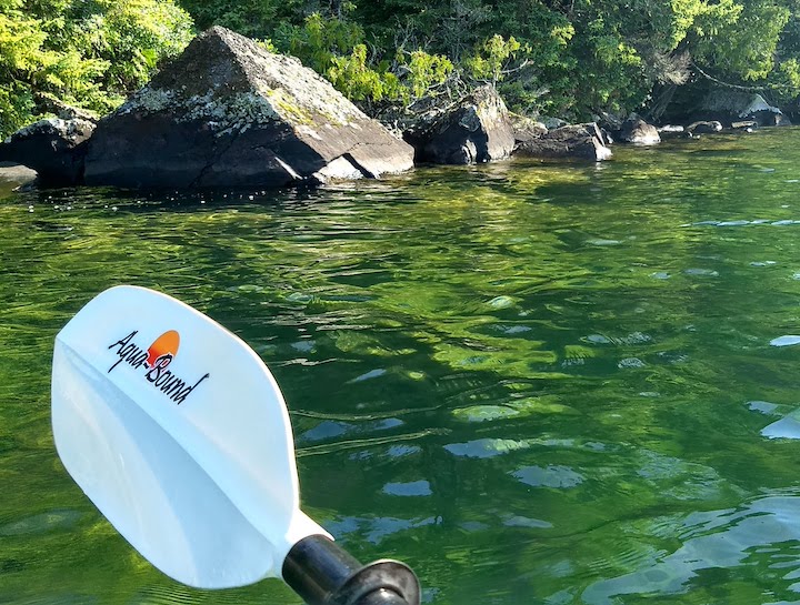 white kayak paddle blade over green water, rocky shoreline