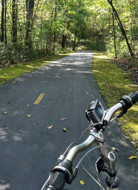 looking over bike's handlebars onto the paved trail through the woods