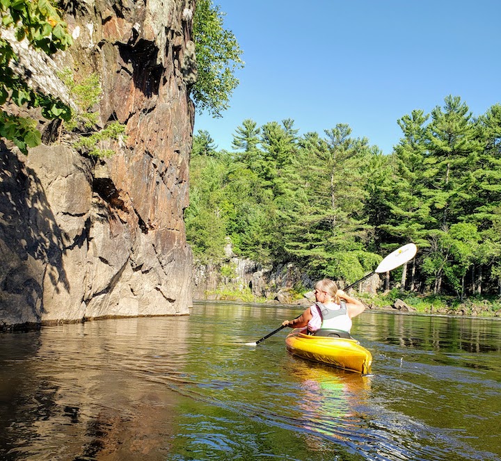 woman kayaking on a river alongside a rocky cliff, with trees on the far bank