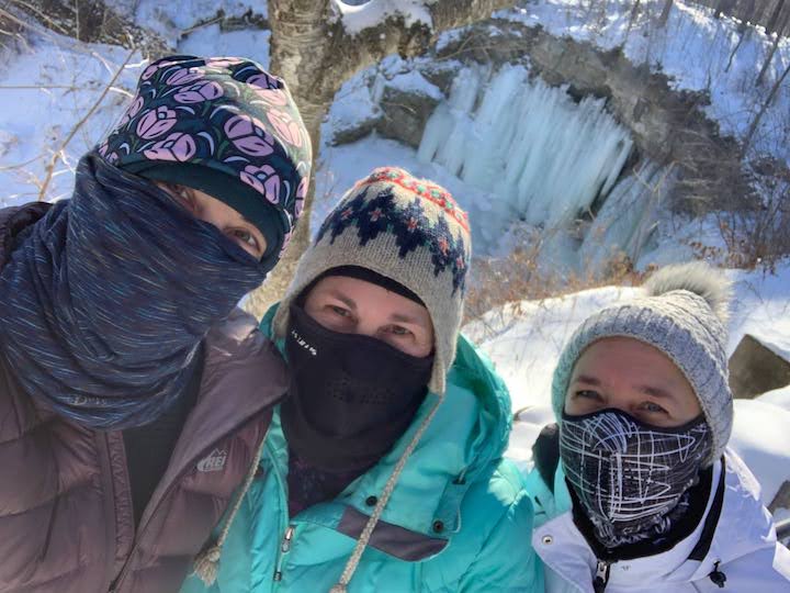 three sisters in winter gear, including face masks; frozen waterfall in the background
