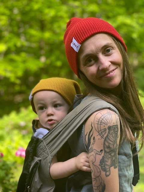 young mom with a red FLORT beanie; baby on her back with a gold FLORT beanie