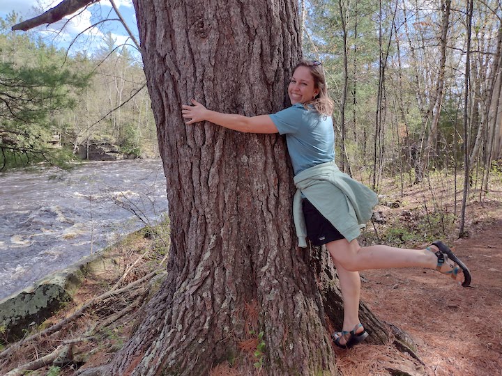 young woman hugging a big tree next to a river, and looking happy