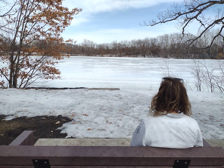 woman sits on a bench and overlooks a still-frozen Silver Lake