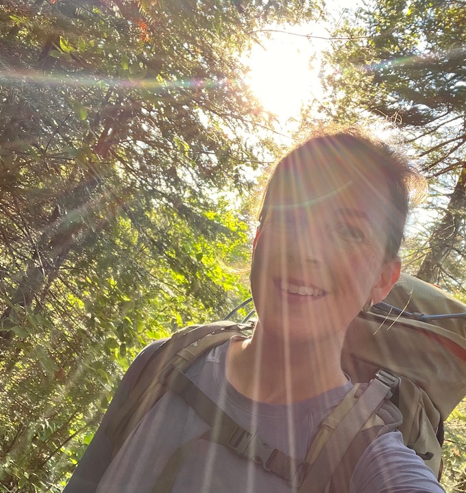woman with a backpack in the woods with the sun streaming in behind her