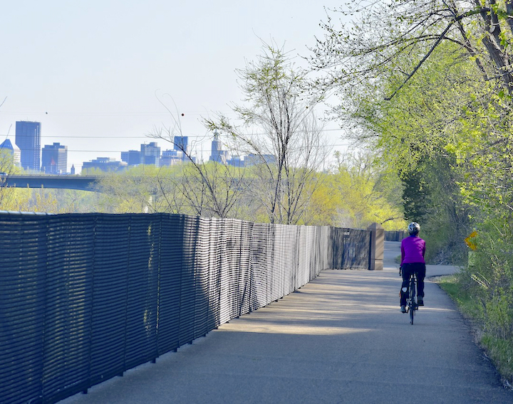 woman bikes on a paved trail with a city skyline in the background