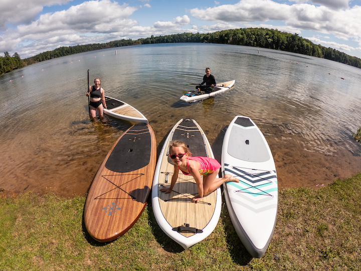 two women and a young girl at a lake shoreline with five Grey Duck paddle boards