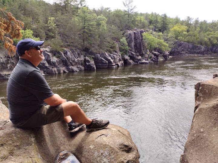 man sits on a rock overlooking the St Croix River, while hiking the trails